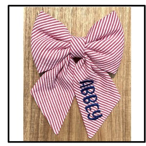 May include: A large, red and white striped bow with the name "ABBEY" embroidered in navy blue. The bow is made of fabric and has a classic bow shape. The background is a light brown wooden surface.