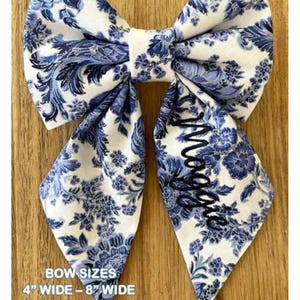 May include: A fabric bow featuring a white and blue floral pattern with the name Maggie embroidered in dark blue thread on the right tail. The text on the image indicates bow sizes ranging from 4 inches to 8 inches wide for S-XXL dogs.