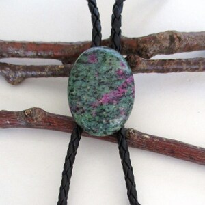 May include: A bolo tie with a green and pink gemstone pendant and black braided leather cords with silver twisted ends.