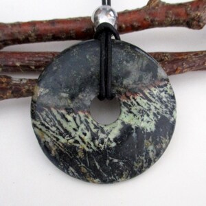 May include: A circular pendant with a central hole, crafted from a stone with a mix of dark green, black, and white patterns. The pendant is suspended from a black cord, with a silver bead accent. The background includes a brown twig.