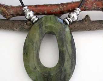 May include: Oval-shaped green pendant necklace with a black cord. The pendant is made of a marbled green stone with a hole in the center. Silver-colored beads accent the cord near the pendant. The necklace is displayed against a natural wood background.