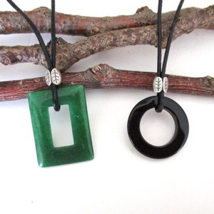 May include: Two necklaces with black leather cords and silver leaf accents. One necklace features a green rectangular pendant with a cutout rectangle in the center. The other necklace features a black circular pendant with a hole in the center.