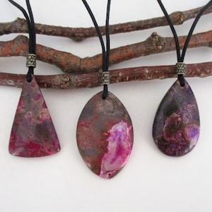May include: Three agate stone pendants on black cords with antique bronze clasps. The stones are pink, purple, and brown with a marbled pattern. The pendants are hanging from a branch.