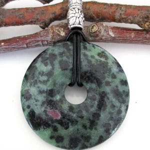 May include: A round, green and black pendant with a central hole, strung on a black cord. The pendant has a speckled pattern and a silver-colored bead. The background includes a brown twig.