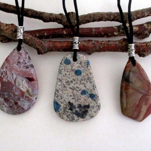 May include: Three teardrop-shaped stone pendants on black cords with silver bead accents. The stones are a mix of red, brown, white, and blue.