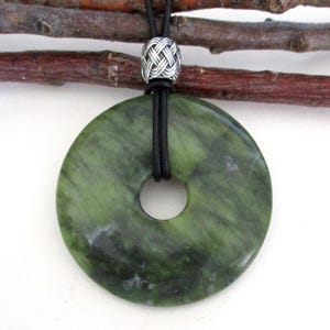 May include: A green jade pendant with a central hole, suspended on a black cord. The cord is held by a silver-colored, intricately woven bead. The jade has a marbled appearance with varying shades of green.