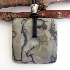 May include: Square pendant necklace with a natural stone. The pendant is a light gray color with black veining and a central hole. It hangs from a black cord with a silver-colored metal bead.