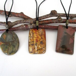 May include: Three stone pendants on black cords with silver and bronze accents. The stones are a green, brown, and red jasper with a marbled pattern.