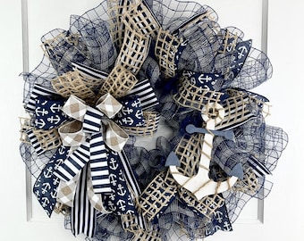 May include: A nautical-themed wreath with navy blue, tan, and white ribbons and mesh. The wreath features a large bow and an anchor accent. The wreath is hanging on a white door.