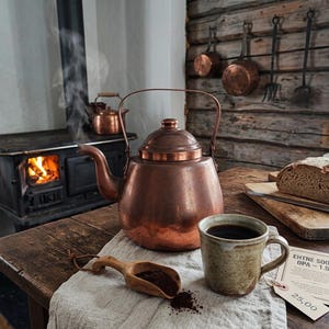 Puede incluir: Una tetera de cobre con un pico humeante, una taza de cerámica llena de líquido oscuro, una cuchara de madera con café molido y una hogaza de pan sobre una tabla de madera. Una etiqueta con el texto EHTNE SOOME OPA - 1.5 L está sobre la mesa.