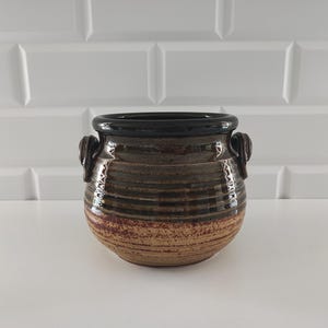 May include: A handcrafted ceramic pot with a dark brown rim and handles. The pot features horizontal bands of brown and tan hues, with a textured, rustic appearance. The interior is dark, and the pot sits on a white surface.
