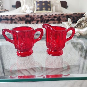 May include: Two ruby red glass serving items, a creamer and a sugar bowl, are on a glass surface. The creamer has a spout and handle, while the sugar bowl has two handles. Both pieces feature a floral design.
