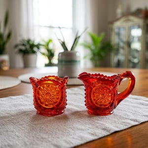Puede incluir: Dos piezas a juego de vidrio rojo anaranjado de estilo vintage se encuentran sobre un mantel de lino gris claro. Uno es un jarrito con asa y boquilla, el otro un pequeño recipiente cuadrado. Ambos tienen un diseño floral.