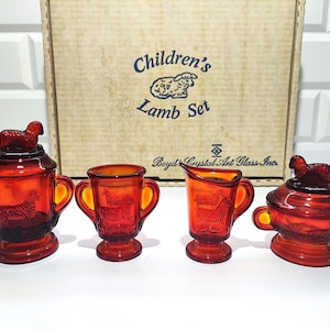 Puede incluir: Un juego de cordero para niños vintage de cristal ámbar. El juego incluye un recipiente con tapa y cordero, dos tazas con asas y una jarra de crema. La caja detrás del juego dice "Children's Lamb Set".