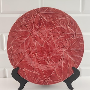 以下が含まれることがあります： 赤い丸い装飾皿で、浮き彫りの葉のデザインが特徴です。 黒いスタンドで支えられており、秋や自然をテーマにしています。 皿の表面は、詳細な葉の印象で覆われています。