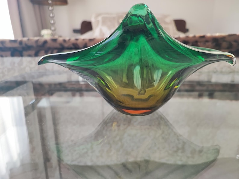 May include: A decorative glass bowl with a unique, organic shape. The bowl transitions from a deep green at the top to a golden yellow at the base. Its form is reminiscent of a flower or a stylized leaf, adding a touch of elegance to any space.