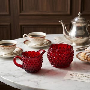 Op de afbeelding: Een stilleven met een robijnrode glazen roomkannetje en een kom met hobnail-patroon. Twee kopjes met schotels, een zilveren theepot en croissants zijn ook aanwezig. De items zijn op een marmeren tafel geplaatst.