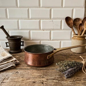 May include: A copper saucepan with a long brass handle and a silver-toned interior lining. The pan sits on a wooden surface alongside a stack of folded linen fabric, a dark metal mortar and pestle, a ceramic jar with wooden spoons, and herbs.