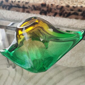 May include: A decorative glass bowl with a unique shape and a gradient color scheme. The bowl transitions from yellow to green, creating a vibrant and eye-catching piece. The bowl is sitting on a glass table.
