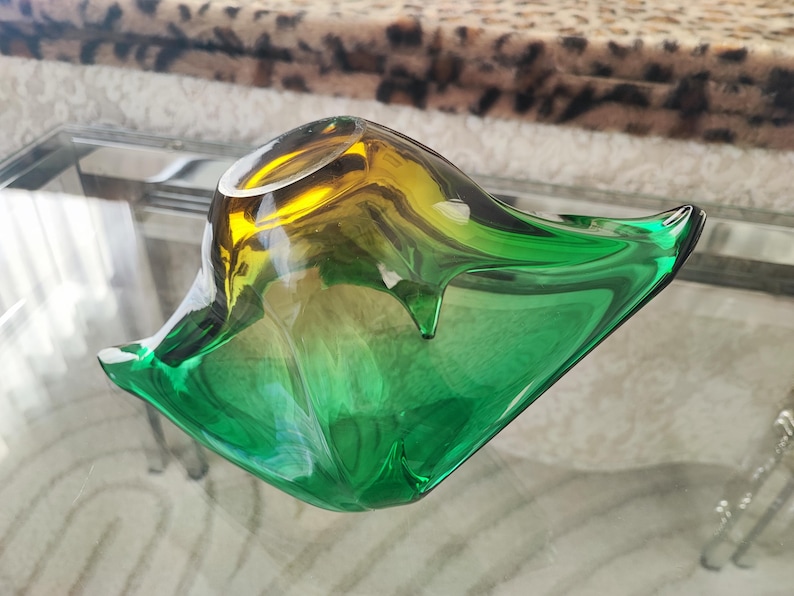 May include: A decorative glass bowl with a unique shape and a gradient color scheme. The bowl transitions from yellow to green, creating a vibrant and eye-catching piece. The bowl is sitting on a glass table.