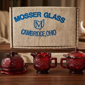 May include: A trio of ruby red glass serving dishes with an intricate pattern. The set comprises a covered dish, a creamer, and a sugar bowl. A cardboard box with "MOSSER GLASS CAMBRIDGE, OHIO" is in the background.