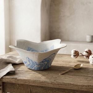 May include: A white ceramic bowl with a flared, petal-like rim and a blue marbled pattern. The bowl features a smooth finish and a wide opening, showcasing a distinct wavy design that wraps around the exterior and interior surfaces.