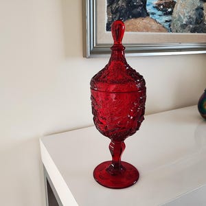 Könnte beinhalten: Ein hoher, rubinroter Glasbehälter mit Deckel und einem verzierten, strukturierten Muster. Der Behälter steht auf einem Sockel und hat einen Griff am Deckel. Das dekorative Stück steht auf einer weißen Oberfläche.