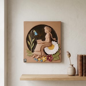 Puede incluir: Una pieza de arte mural cuadrada de cerámica con una escultura en relieve de una mujer leyendo. La obra de arte incluye una campanilla, una gran margarita y otras flores coloridas. La pieza está sobre un fondo neutro.