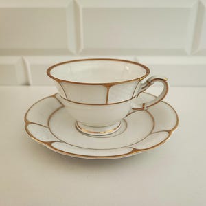Può includere: Un set di tazza e piattino bianchi con bordi dorati. La tazza ha una forma arrotondata con un manico, e il piattino ha un bordo smerlato. I bordi dorati delineano la tazza e il piattino, aggiungendo un tocco di eleganza.