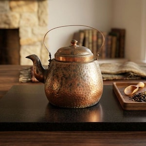 May include: A copper tea kettle with a hammered texture sits on a black surface. A wooden tray holds a glass of tea, tea leaves, and a small wooden spoon. The kettle has a curved spout and a handle.