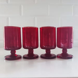 Puede incluir: Un juego de cuatro copas de cristal rojo rubí a juego, cada una con un cuenco cilíndrico con estrías verticales, un tallo corto y grueso, y una base circular. La cristalería es translúcida y de color uniforme, dispuesta en fila.