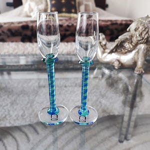 Puede incluir: Dos copas de champán transparentes con tallos y bases en espiral azul verdoso y azul. Los tallos tienen un diseño retorcido. Las copas están sobre una mesa de cristal, con una figura decorativa en el fondo.
