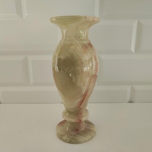 Pu&ograve; includere: Vaso decorativo in onice, con un motivo venato beige chiaro e rosso. Il vaso ha una parte superiore svasata, un corpo bulboso e una base rotonda. Altezza di circa 20 cm.
