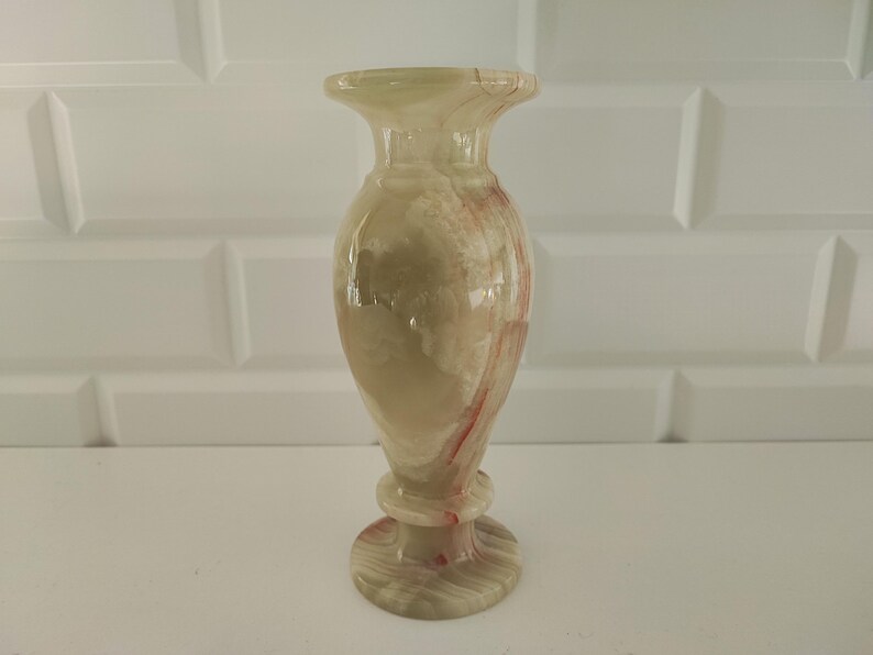 Pu&ograve; includere: Vaso decorativo in onice, con un motivo venato beige chiaro e rosso. Il vaso ha una parte superiore svasata, un corpo bulboso e una base rotonda. Altezza di circa 20 cm.
