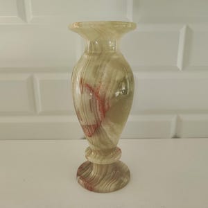Pu&ograve; includere: Vaso decorativo in onice, con parte superiore svasata e base arrotondata. Il vaso presenta una palette di colori naturali beige, marrone e rosso, con finitura lucida e brillante. Il vaso &egrave; alto circa 20 cm.