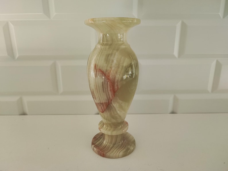 Pu&ograve; includere: Vaso decorativo in onice, con parte superiore svasata e base arrotondata. Il vaso presenta una palette di colori naturali beige, marrone e rosso, con finitura lucida e brillante. Il vaso &egrave; alto circa 20 cm.