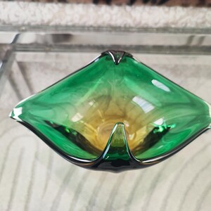 May include: A green and yellow glass ashtray with a unique, geometric shape. The ashtray has a triangular form with rounded edges and a small, pointed lip. The glass has a gradient effect, transitioning from green to yellow. The ashtray is a decorative item.