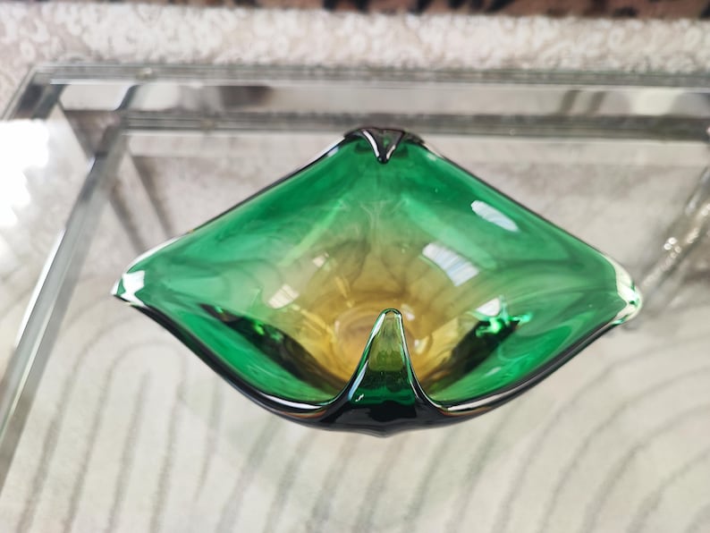 May include: A green and yellow glass ashtray with a unique, geometric shape. The ashtray has a triangular form with rounded edges and a small, pointed lip. The glass has a gradient effect, transitioning from green to yellow. The ashtray is a decorative item.