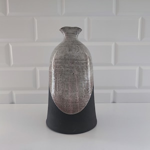 Może przedstawiać: Ręcznie wykonany ceramiczny wazon o unikalnym designie. Górna część jest nakrapiana na szaro, przechodząc w czerwonobrązowy odcień, a podstawa jest czarna. Wazon ma wąską szyjkę i zaokrąglony korpus, o wysokości około 25 cm.