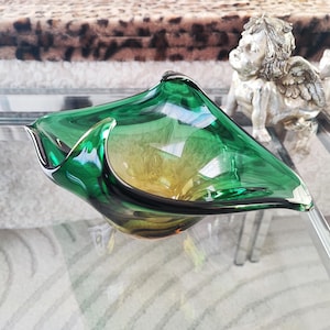 May include: A green and yellow glass bowl with a unique, asymmetrical shape. The bowl has a glossy finish and sits on a glass table. A small, silver cherub statue is in the background.