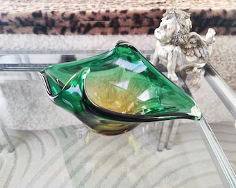 May include: A green and yellow glass bowl with a unique, asymmetrical shape. The bowl has a glossy finish and sits on a glass table. A small, silver cherub statue is in the background.