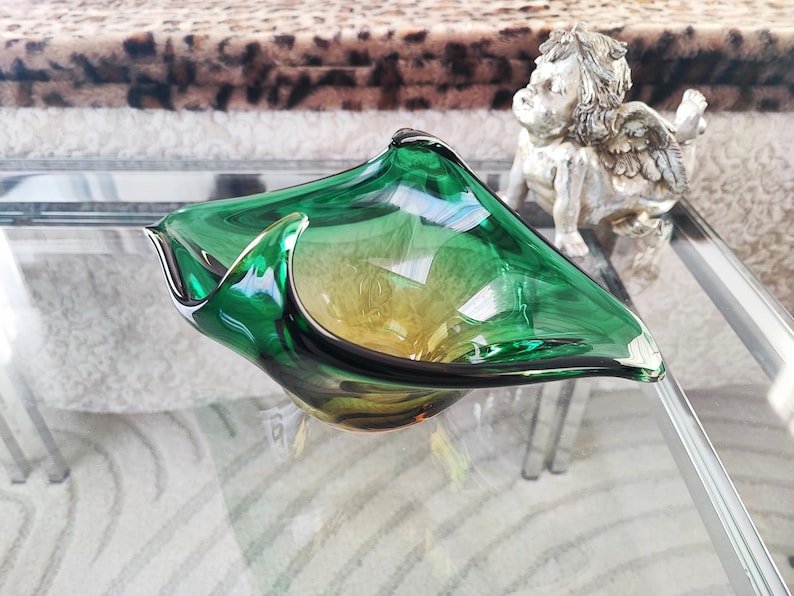 May include: A green and yellow glass bowl with a unique, asymmetrical shape. The bowl has a glossy finish and sits on a glass table. A small, silver cherub statue is in the background.