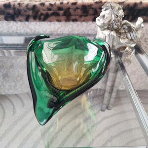May include: A green and yellow glass bowl with a unique, organic shape. The bowl has a glossy finish and is displayed on a glass surface. A small silver angel statue is in the background.