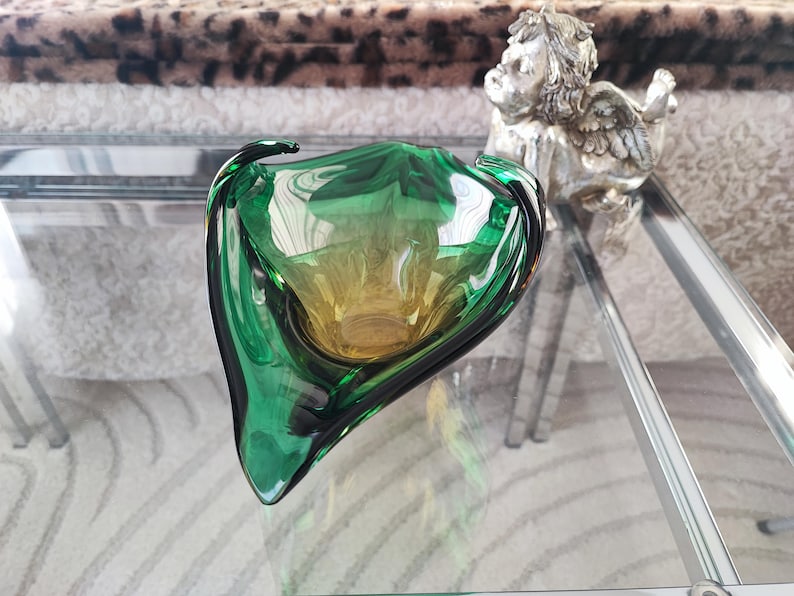May include: A green and yellow glass bowl with a unique, organic shape. The bowl has a glossy finish and is displayed on a glass surface. A small silver angel statue is in the background.