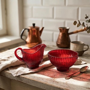 Peut inclure: Un pichet à crème en verre rouge côtelé avec une anse et un bol en verre rouge côtelé assorti. L'ensemble est posé sur un tissu clair. En arrière-plan, on trouve une bouilloire en cuivre, une cafetière en cuivre, une tasse et un vase en verre.