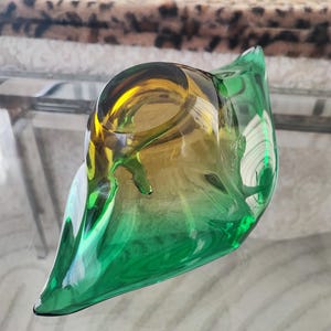 May include: A decorative glass bowl with a unique, organic shape. The bowl transitions from a vibrant green at the base to a golden-yellow hue at the top. Its smooth, curved design adds a touch of elegance to any space.