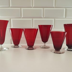May include: A collection of six ruby red glass vessels with flared rims and clear glass bases. The set includes varying heights and widths, each featuring a smooth, translucent red body that tapers down to a circular, transparent glass foot.