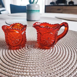 Könnte beinhalten: Zwei antike, leuchtend orangefarbene Sahne- und Zuckerdosen aus Glas. Die Stücke weisen ein Sternmuster und einen gewellten Rand auf. Der Sahnekännchen hat einen Griff. Das Set steht auf einem neutralen Platzset.