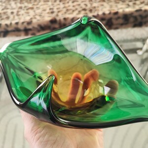 May include: A hand-blown glass bowl with a vibrant green exterior and a warm amber interior. The bowl has a unique, organic shape with pointed edges. The interior features a cluster of orange-brown glass elements, adding depth and visual interest.