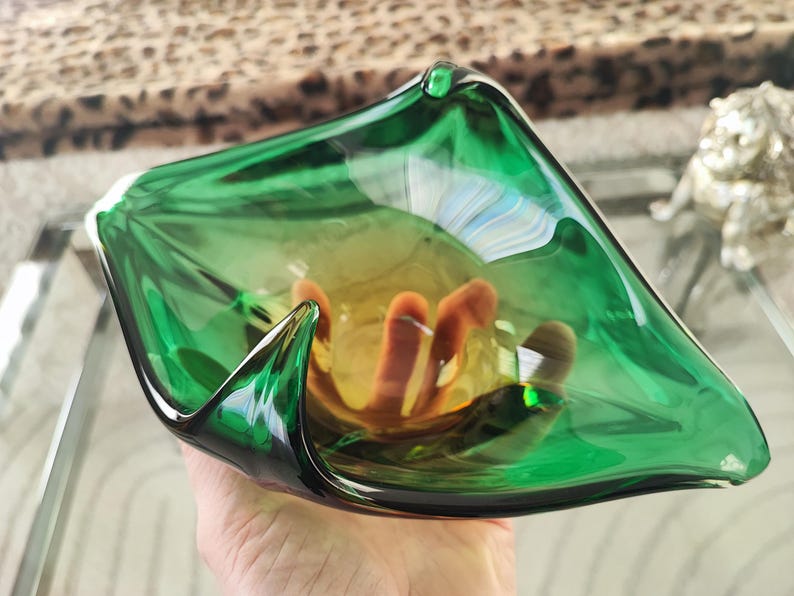 May include: A hand-blown glass bowl with a vibrant green exterior and a warm amber interior. The bowl has a unique, organic shape with pointed edges. The interior features a cluster of orange-brown glass elements, adding depth and visual interest.